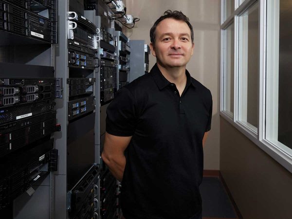 surrounded by racks of computer servers. White-framed windows line the right side of the room. He is smiling and has his hands behind his back.|Smiling woman in a floral blouse stands in a modern office environment with people working at computers in the background. The atmosphere is professional and welcoming.|A person wearing a blue checkered shirt stands smiling in an office environment with cubicles and computers. The background shows other people working at their desks.