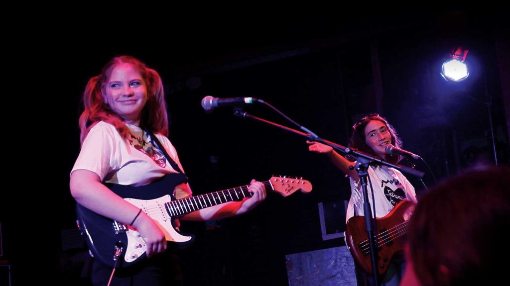 holding a bass guitar. Stage lights illuminate them.|A group of enthusiastic young people sing and raise their hands in a classroom setting. They are led by a person with a microphone. Musical instruments like a keyboard and a guitar are visible. Posters with messages adorn the walls.|A group of people sit in a circle in a classroom setting. They are engaged in conversation. Instruments are visible in the background