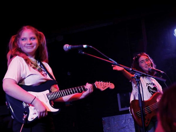 holding a bass guitar. Stage lights illuminate them.|A group of enthusiastic young people sing and raise their hands in a classroom setting. They are led by a person with a microphone. Musical instruments like a keyboard and a guitar are visible. Posters with messages adorn the walls.|A group of people sit in a circle in a classroom setting. They are engaged in conversation. Instruments are visible in the background