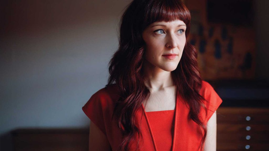 wavy red hair and bangs is wearing a red outfit. She is gazing thoughtfully to the side