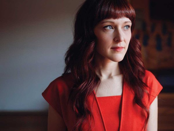 wavy red hair and bangs is wearing a red outfit. She is gazing thoughtfully to the side