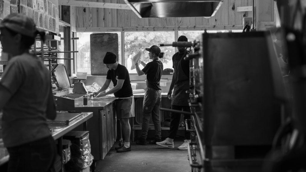 Inside the Shakespeare's pizza shop. 3 people are seen making pizzas in black and white.